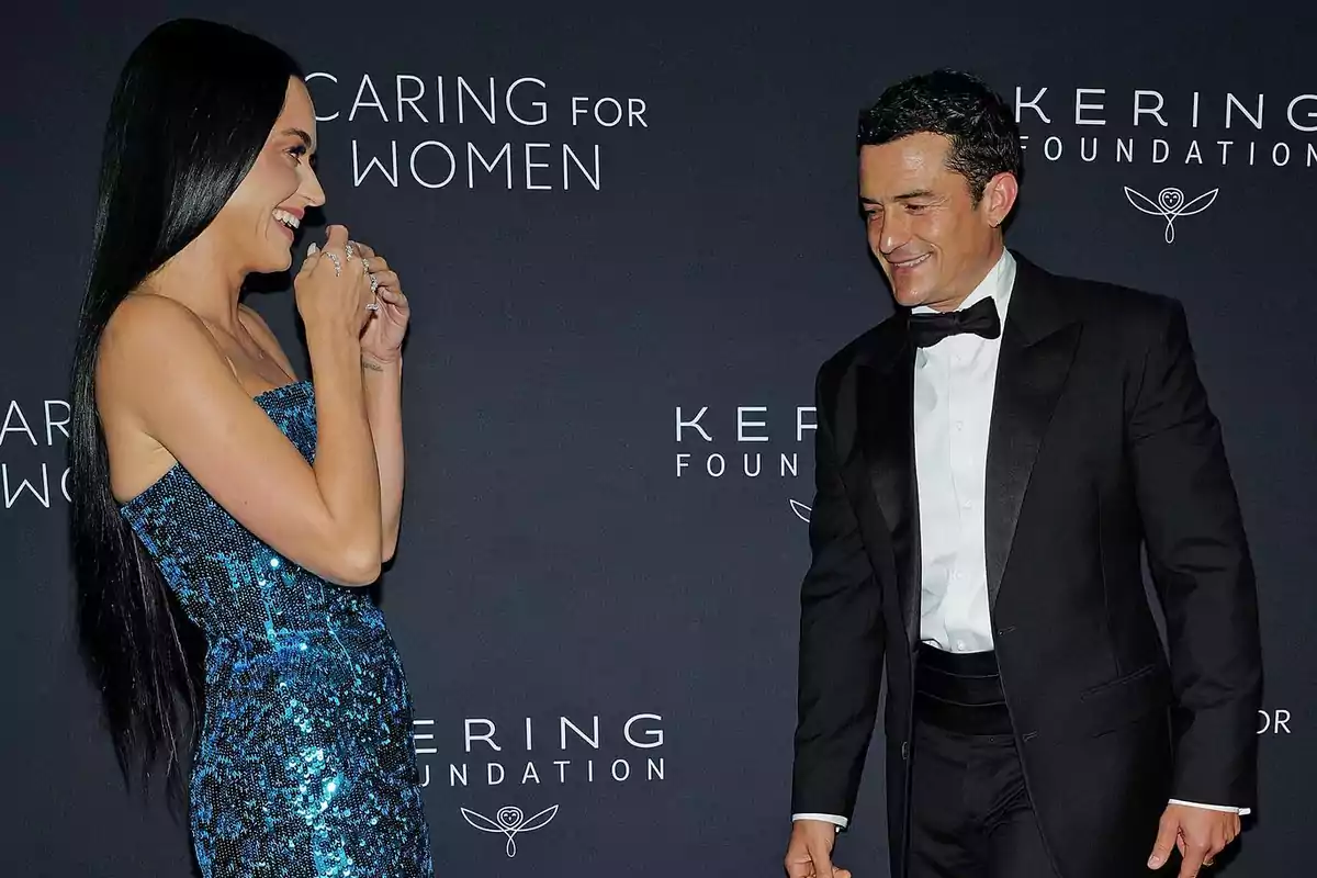 Una mujer con vestido azul brillante y un hombre con esmoquin negro sonríen mientras posan juntos en un evento elegante con fondo oscuro