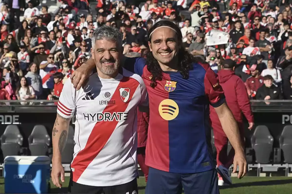 Dos hombres sonríen abrazados en una cancha de fútbol, uno lleva la camiseta de River Plate y el otro la del Barcelona, con público en las gradas al fondo.