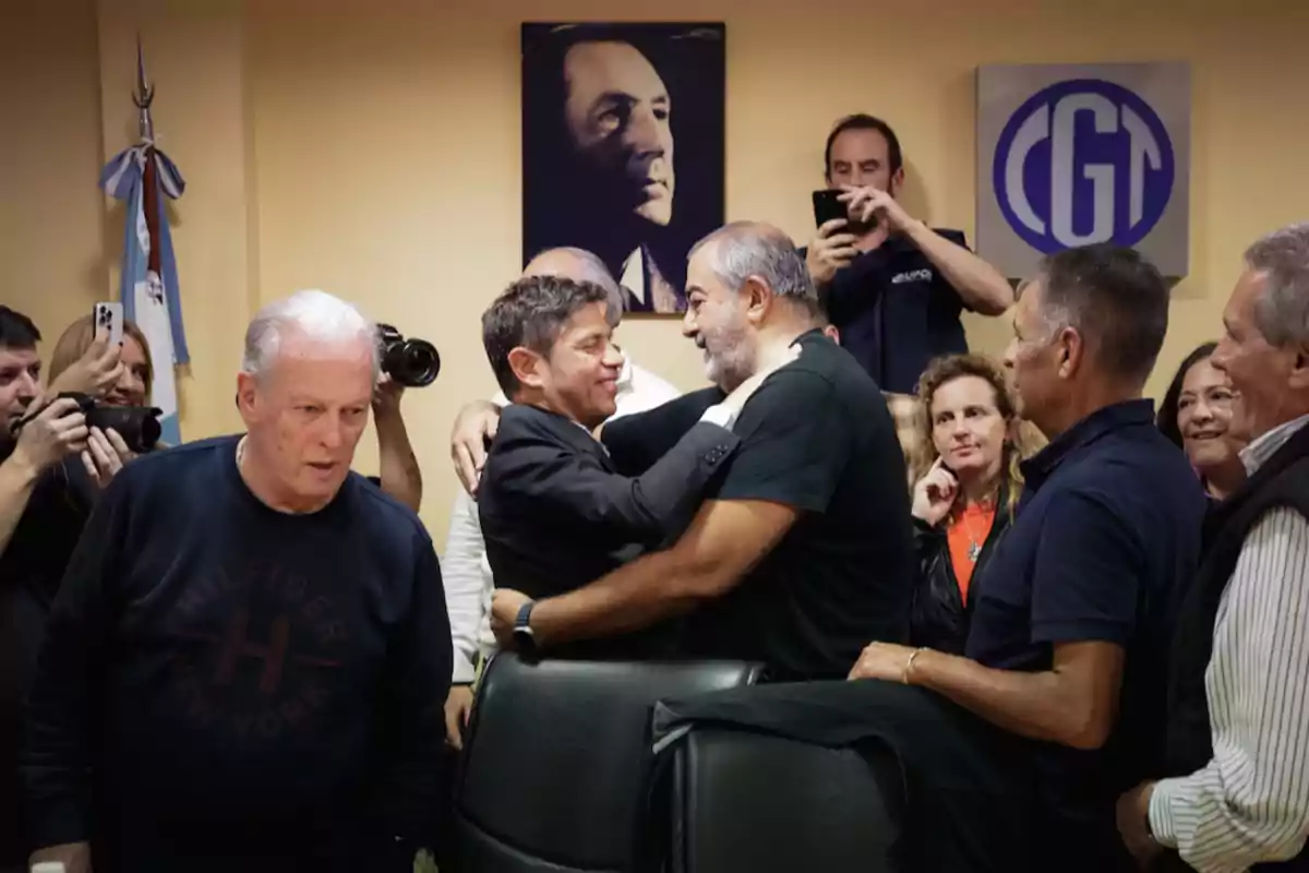 A group of people at a meeting smile and hug while some photographers capture the moment in a room with an Argentine flag and a portrait on the wall.
