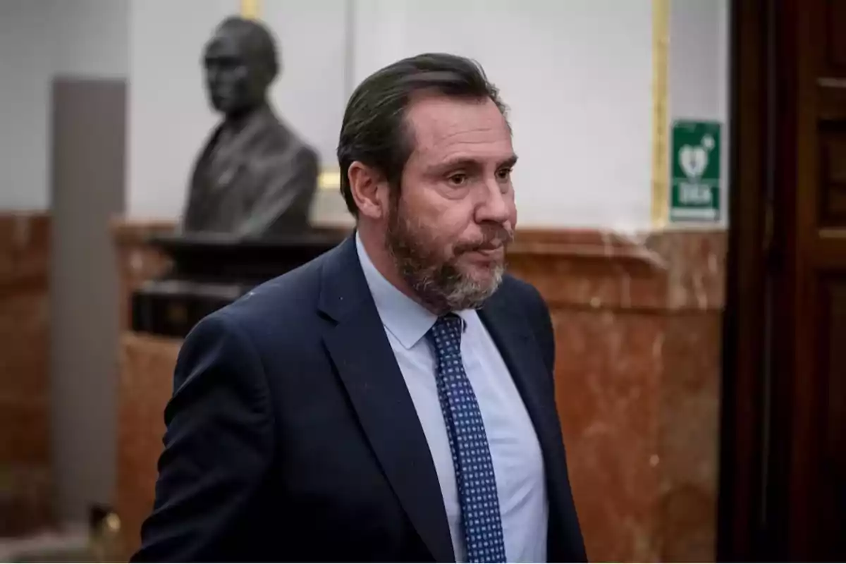 Hombre de traje y corbata azul caminando en un edificio con paredes de mármol y una escultura de busto al fondo