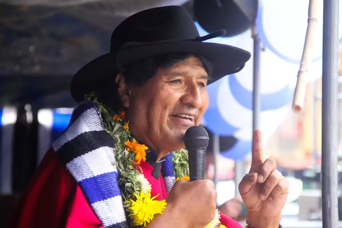 Hombre con sombrero negro y guirnalda de flores hablando por micrófono y levantando un dedo en un evento público