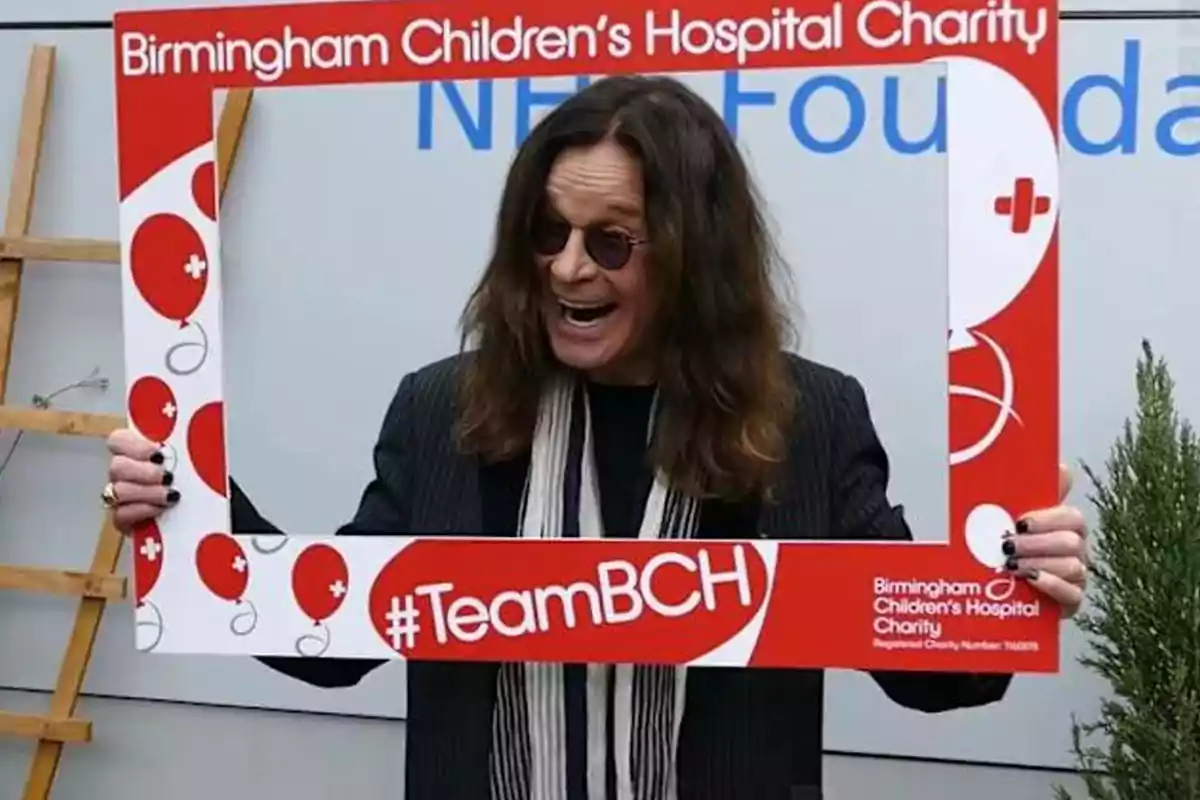 Person with dark glasses and long hair smiles while holding a decorative frame from the Birmingham Children’s Hospital Charity organization with red balloons and the hashtag TeamBCH. Person with dark glasses and long hair smiles while holding a decorative frame from the Birmingham Children’s Hospital Charity organization with red balloons and the hashtag TeamBCH.