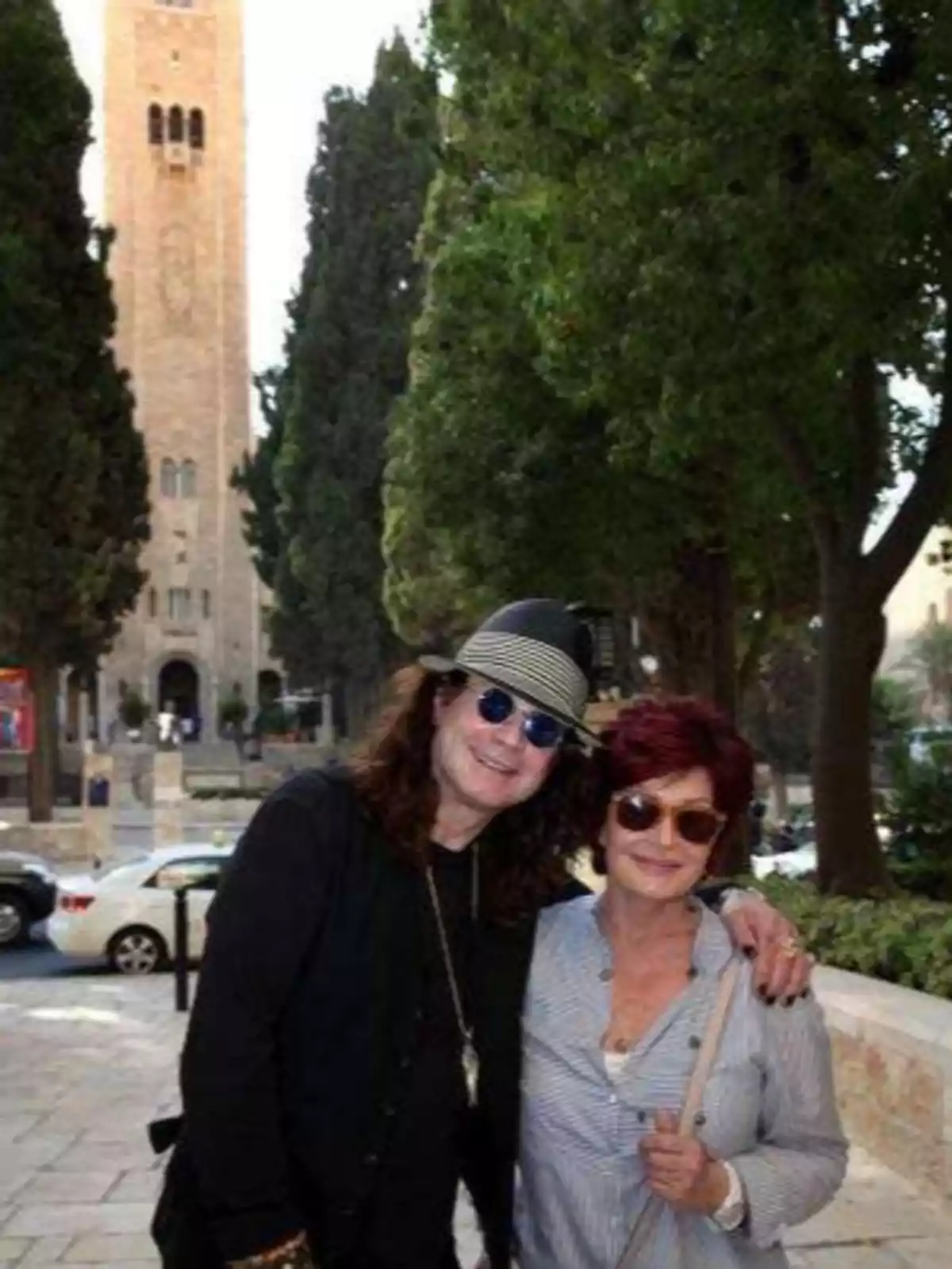 Dos personas sonríen y posan juntas en una calle arbolada con una torre de piedra al fondo Dos personas sonríen y posan juntas en una calle arbolada con una torre de piedra al fondo