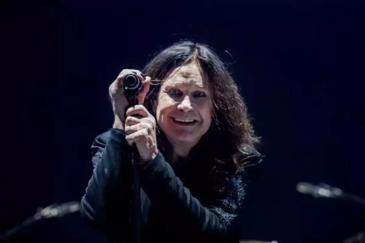 Persona con cabello largo y oscuro sonríe mientras sostiene un micrófono en un escenario con fondo oscuro