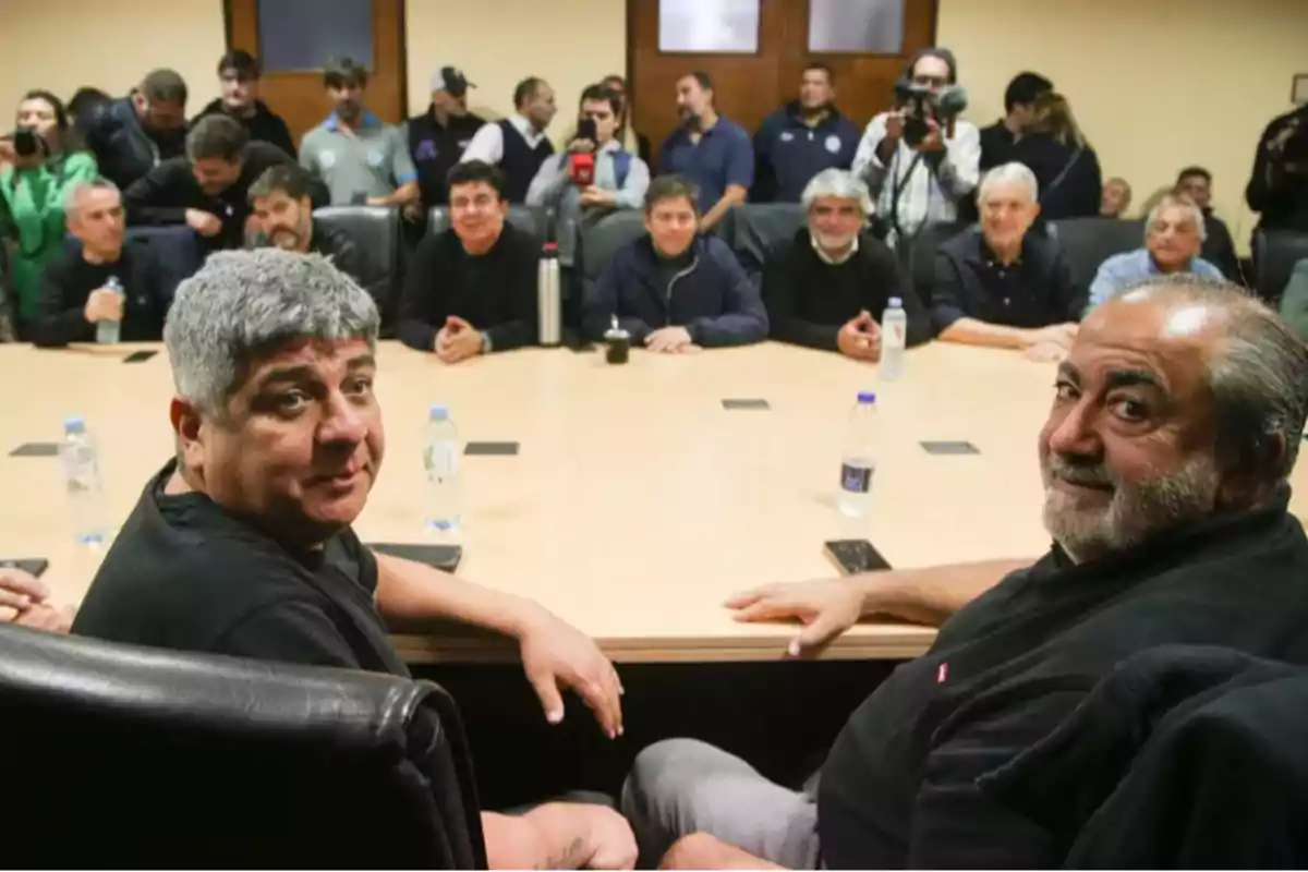 Meeting of several people seated around a large table in a conference room, with two men in the foreground looking toward the camera and several water bottles on the table. Meeting of several people seated around a large table in a conference room, with two men in the foreground looking toward the camera and several water bottles on the table.