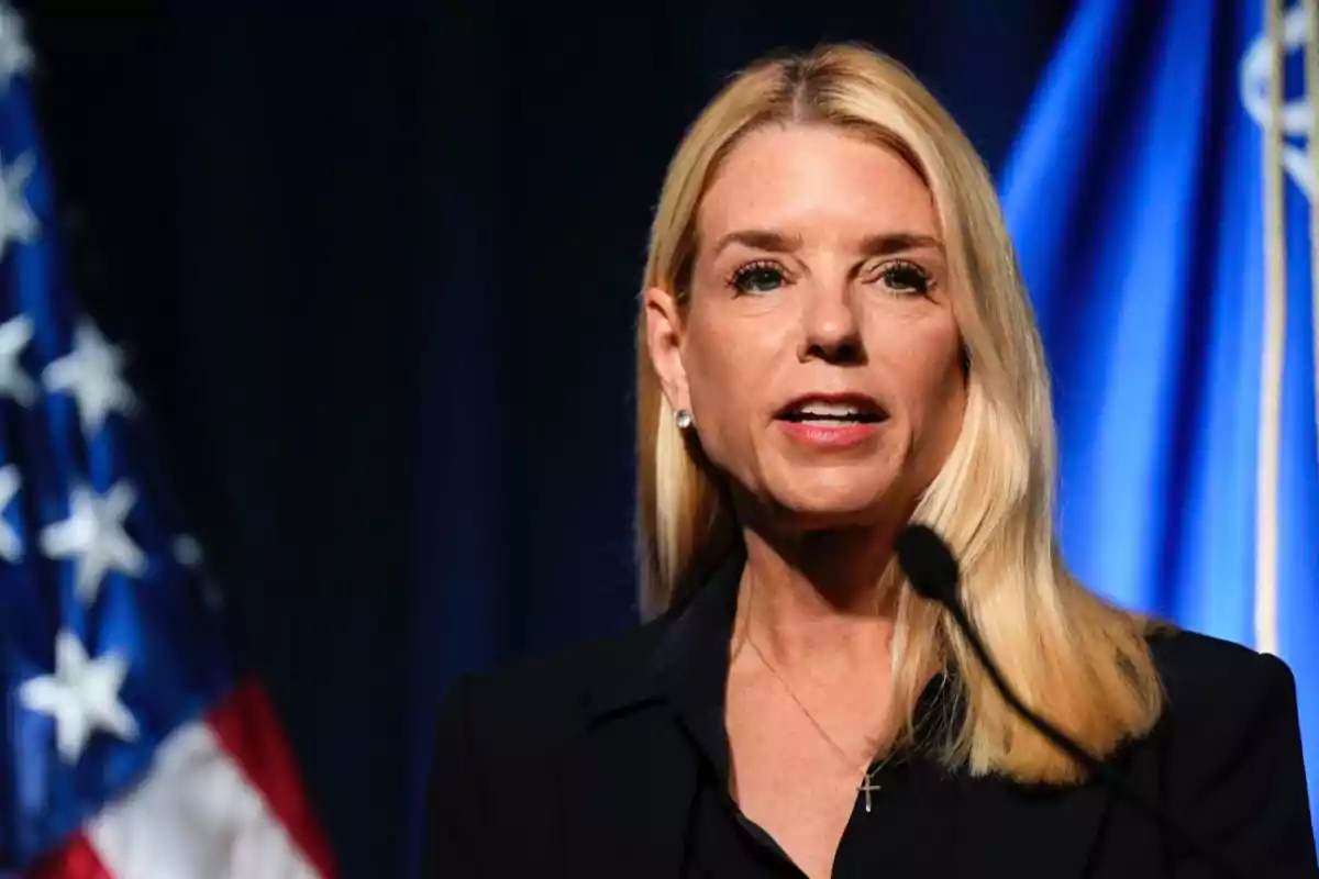 Blonde woman speaking in front of a microphone with a United States flag in the background