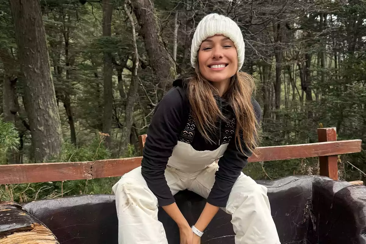 Smiling young woman wearing a white knit hat and winter clothes sitting outdoors in a wooded setting