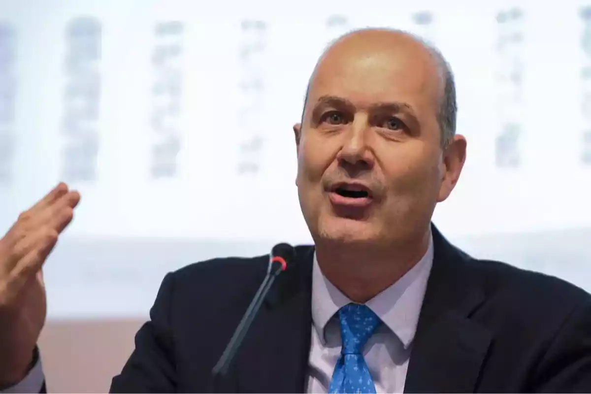 Middle-aged bald man in a suit and blue tie speaking in front of a microphone and gesturing with his hand Middle-aged bald man in a suit and blue tie speaking in front of a microphone and gesturing with his hand