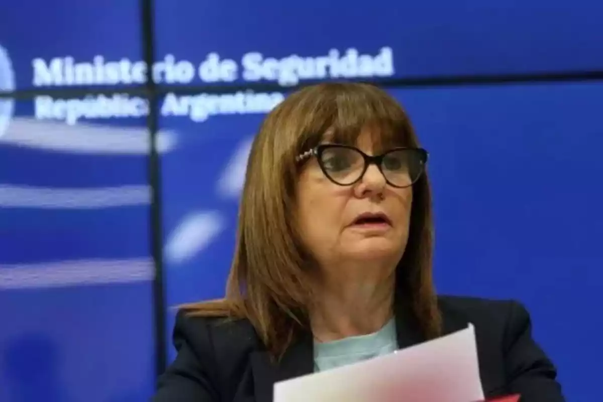 Mujer de cabello castaño y gafas hablando frente a un fondo azul con texto borroso Mujer de cabello castaño y gafas hablando frente a un fondo azul con texto borroso