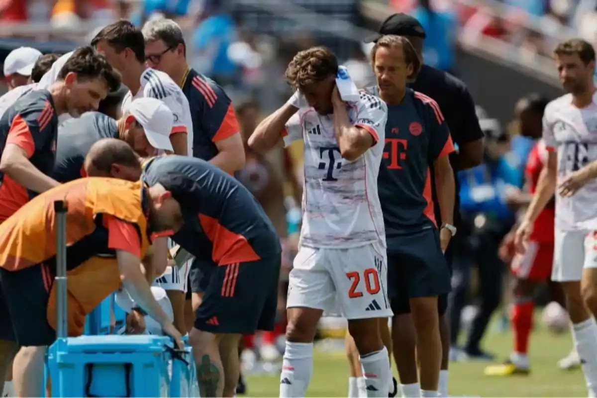Un futbolista con el número 20 en su uniforme blanco se seca el sudor con una toalla mientras varios miembros del equipo técnico y jugadores se agrupan a su alrededor en el campo durante un partido bajo el sol Un futbolista con el número 20 en su uniforme blanco se seca el sudor con una toalla mientras varios miembros del equipo técnico y jugadores se agrupan a su alrededor en el campo durante un partido bajo el sol