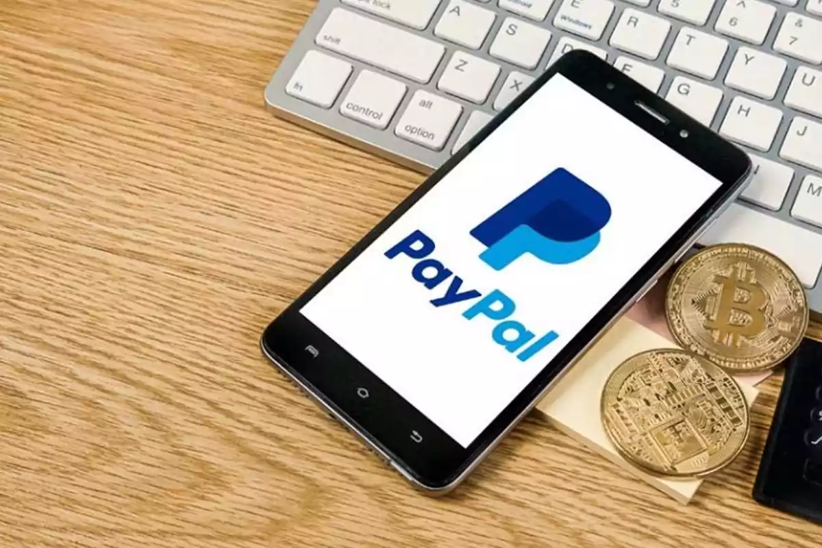 Teléfono móvil con el logo de PayPal en la pantalla junto a dos monedas de bitcoin sobre un escritorio de madera y un teclado de computadora. Teléfono móvil con el logo de PayPal en la pantalla junto a dos monedas de bitcoin sobre un escritorio de madera y un teclado de computadora.