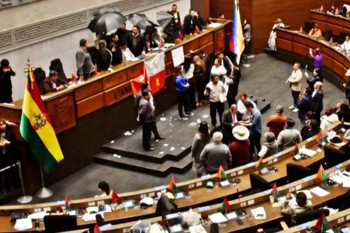 People gathered inside a parliament with Bolivian and Wiphala flags, some people standing and others sitting, while papers can be seen on the floor and there is an atmosphere of discussion or protest.