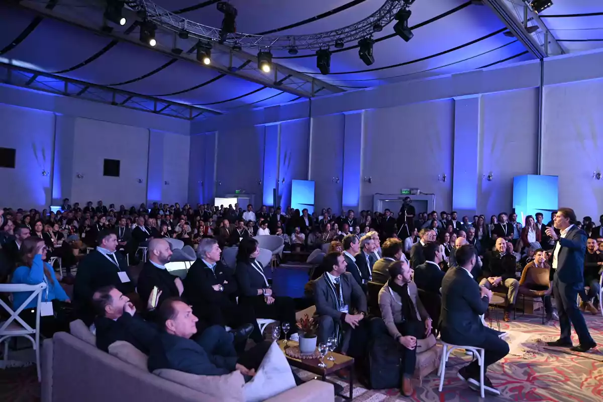 A man standing gives a lecture before a large audience in a hall lit with blue lights.