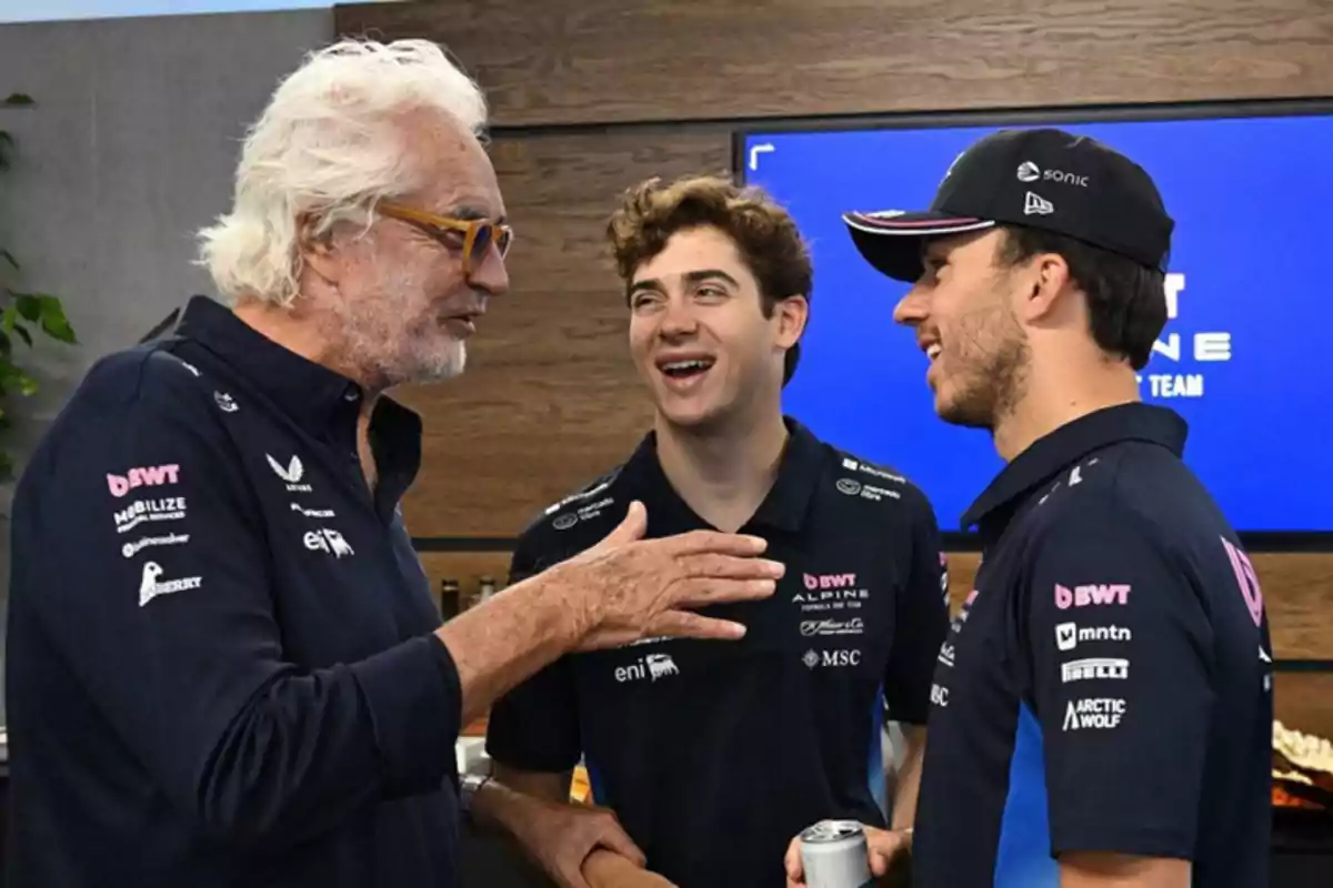 Three men wearing Alpine team clothing talk and smile indoors in front of a blue screen. Three men wearing Alpine team clothing talk and smile indoors in front of a blue screen.