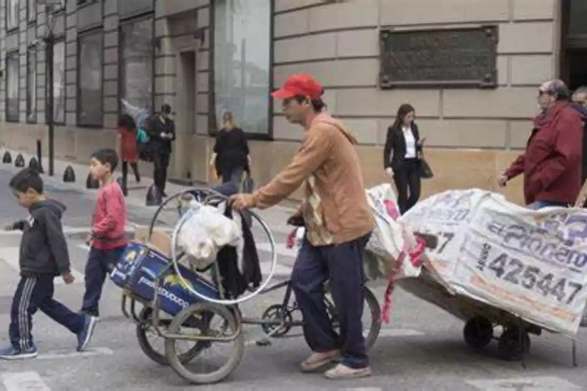 Un hombre empuja un carrito cargado con bolsas grandes mientras dos niños caminan delante de él por una calle urbana con varias personas al fondo Un hombre empuja un carrito cargado con bolsas grandes mientras dos niños caminan delante de él por una calle urbana con varias personas al fondo