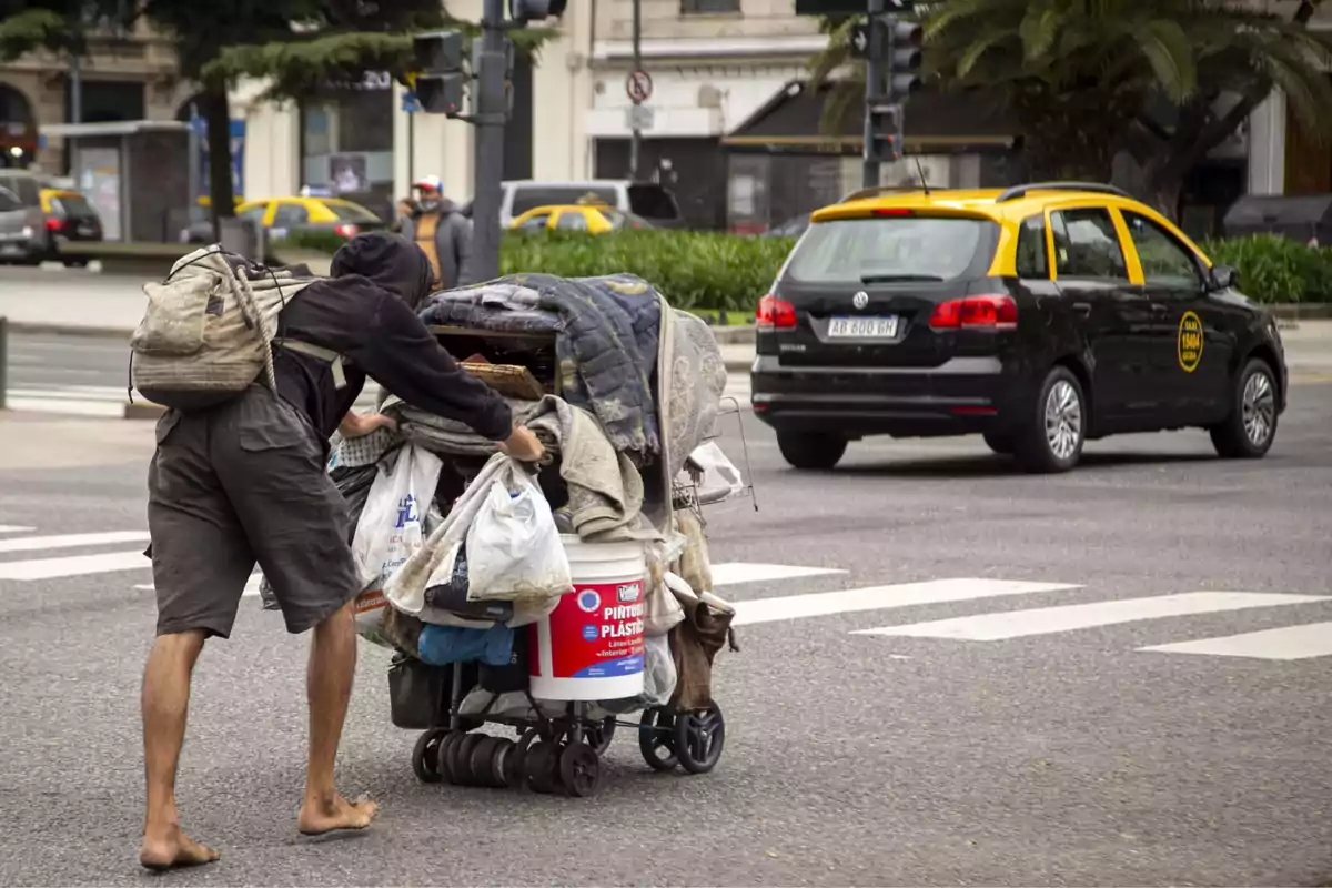 Persona descalza empujando un carrito lleno de pertenencias por una calle urbana mientras un taxi pasa cerca.