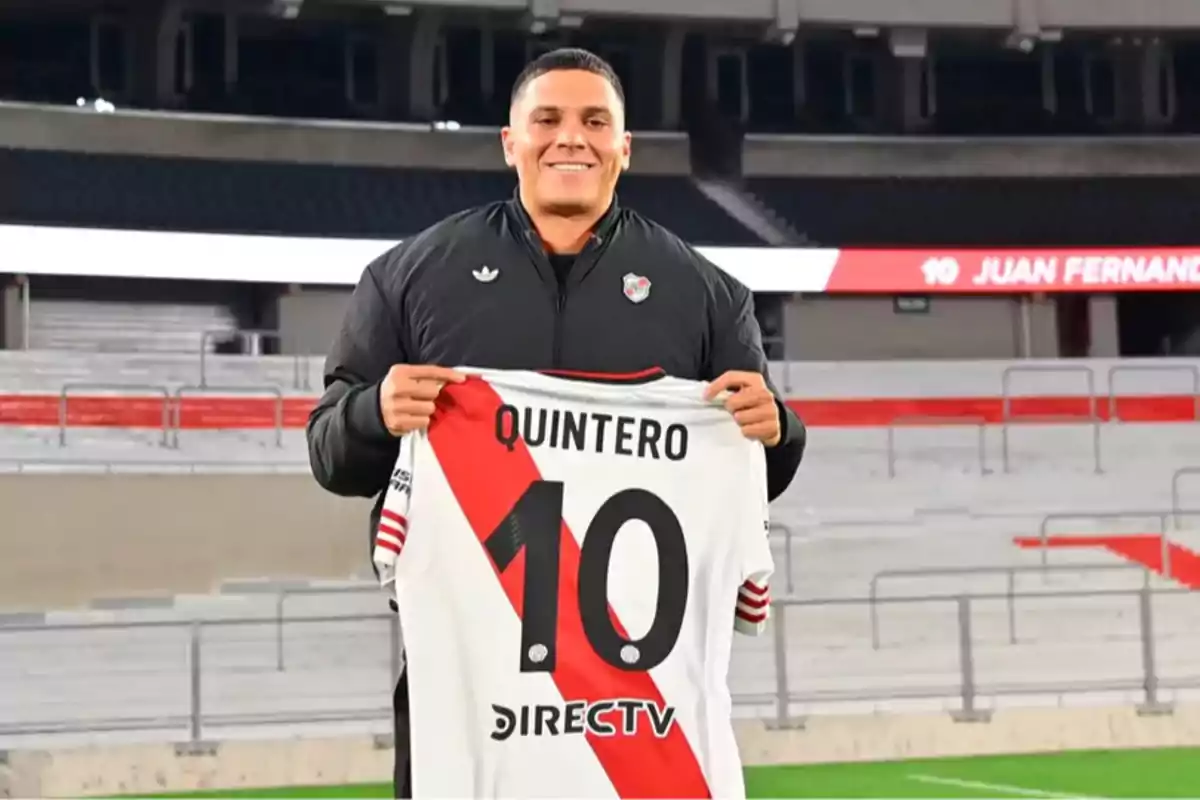 Un hombre sonriente sostiene una camiseta de fútbol blanca con una franja roja y el número 10 en un estadio vacío