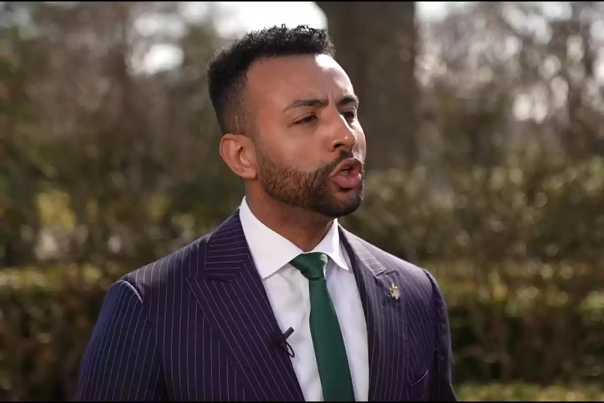 Man with a beard and short hair wearing a pinstripe suit and green tie speaking outdoors Man with a beard and short hair wearing a pinstripe suit and green tie speaking outdoors