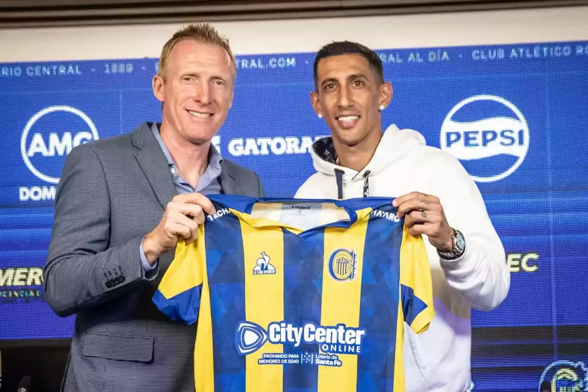 Two people smile while holding a Rosario Central soccer jersey with a background of advertising signs.