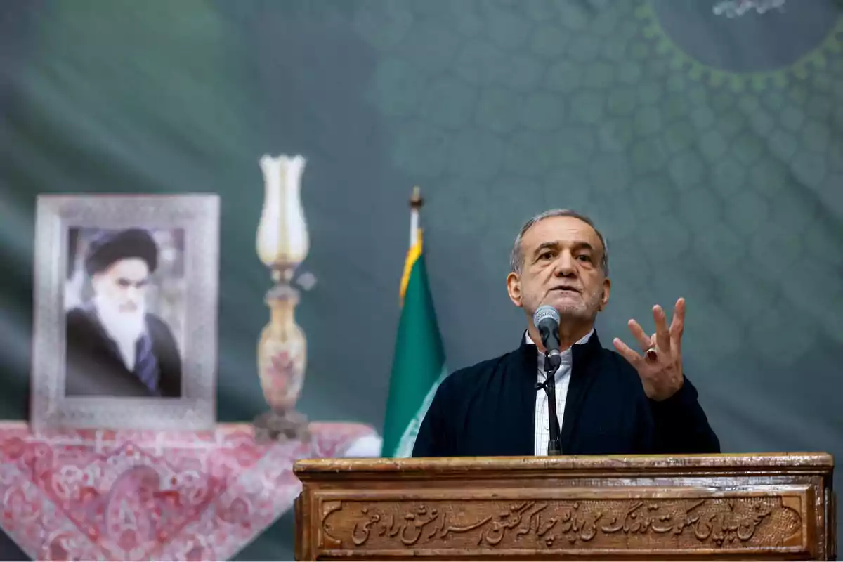 Older man speaking at a podium with a microphone, in the background a flag, a framed photograph, and a decorative lamp on a table covered with a red tablecloth