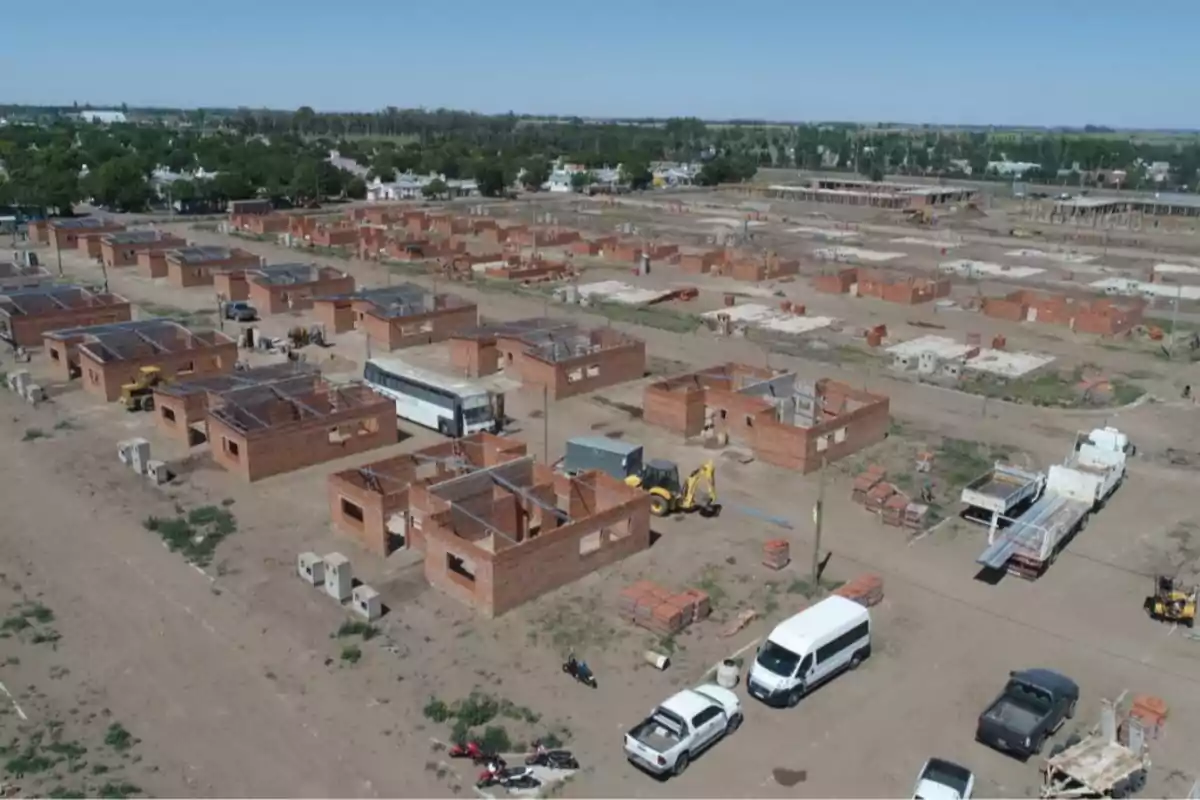 Vista aérea de un conjunto de casas en construcción con paredes de ladrillo, maquinaria y vehículos estacionados en un terreno amplio