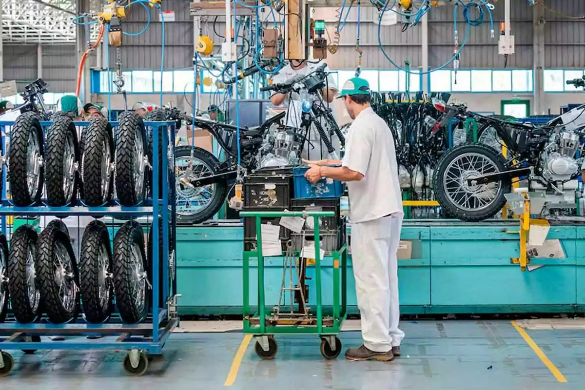 Trabajador ensamblando motocicletas en una línea de producción dentro de una fábrica con varias llantas apiladas en un estante azul Trabajador ensamblando motocicletas en una línea de producción dentro de una fábrica con varias llantas apiladas en un estante azul