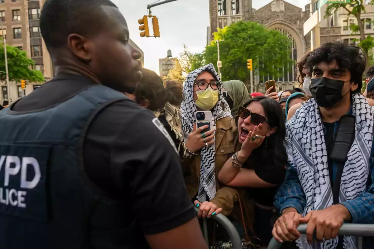 A group of protesters wearing keffiyehs and masks confronts a police officer with his back turned during a street protest; a woman is shouting while another holds a cell phone, and several people watch the scene.