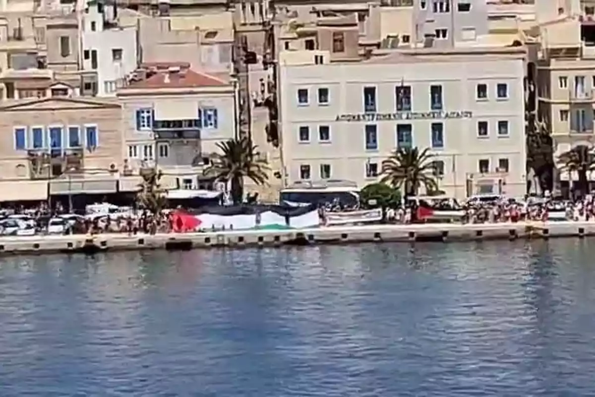 Personas sostienen una gran bandera de Palestina en un muelle frente a edificios antiguos junto al mar Personas sostienen una gran bandera de Palestina en un muelle frente a edificios antiguos junto al mar
