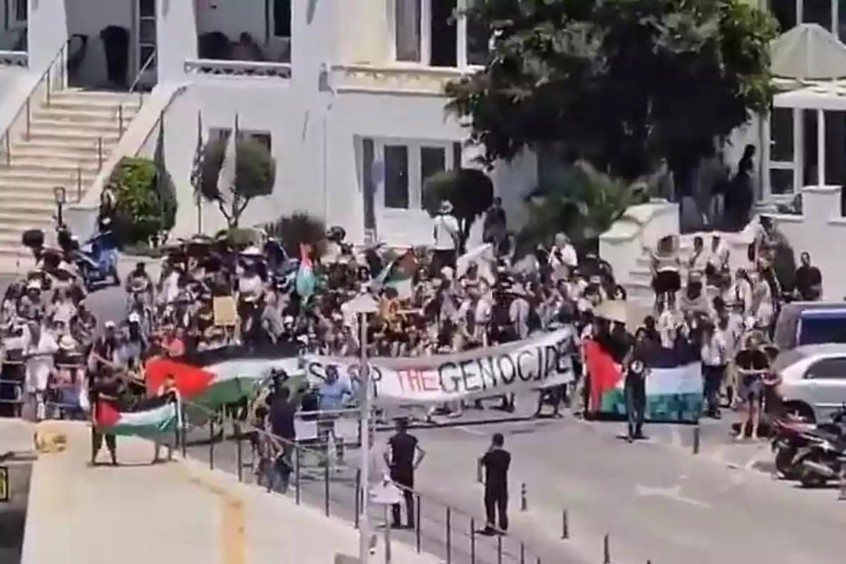 Manifestación frente a un edificio blanco con personas sosteniendo pancartas y banderas palestinas