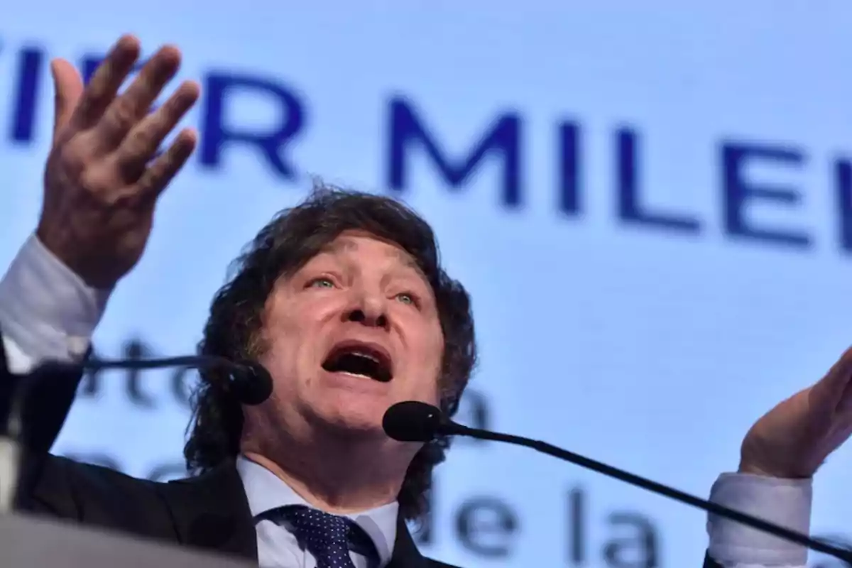 Hombre de cabello castaño y traje oscuro hablando apasionadamente frente a un micrófono con un fondo azul y letras grandes desenfocadas Hombre de cabello castaño y traje oscuro hablando apasionadamente frente a un micrófono con un fondo azul y letras grandes desenfocadas