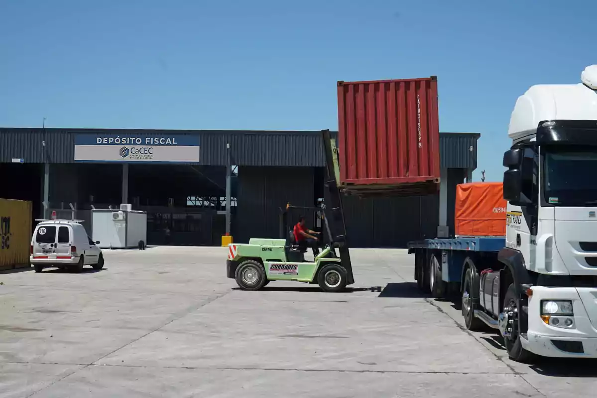Un montacargas levanta un contenedor rojo para colocarlo sobre un camión frente a un depósito fiscal en un área industrial Un montacargas levanta un contenedor rojo para colocarlo sobre un camión frente a un depósito fiscal en un área industrial