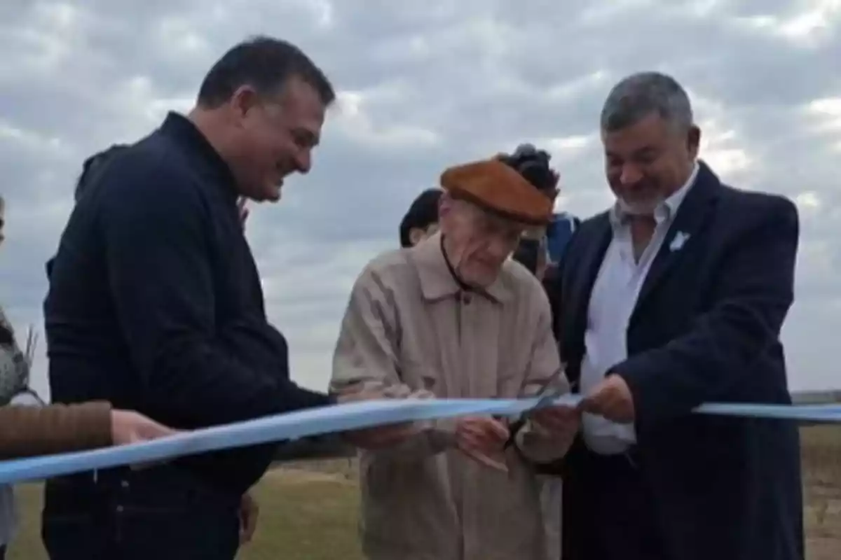 Three men smile as they cut a light blue ribbon at an outdoor event under a cloudy sky. Three men smile as they cut a light blue ribbon at an outdoor event under a cloudy sky.