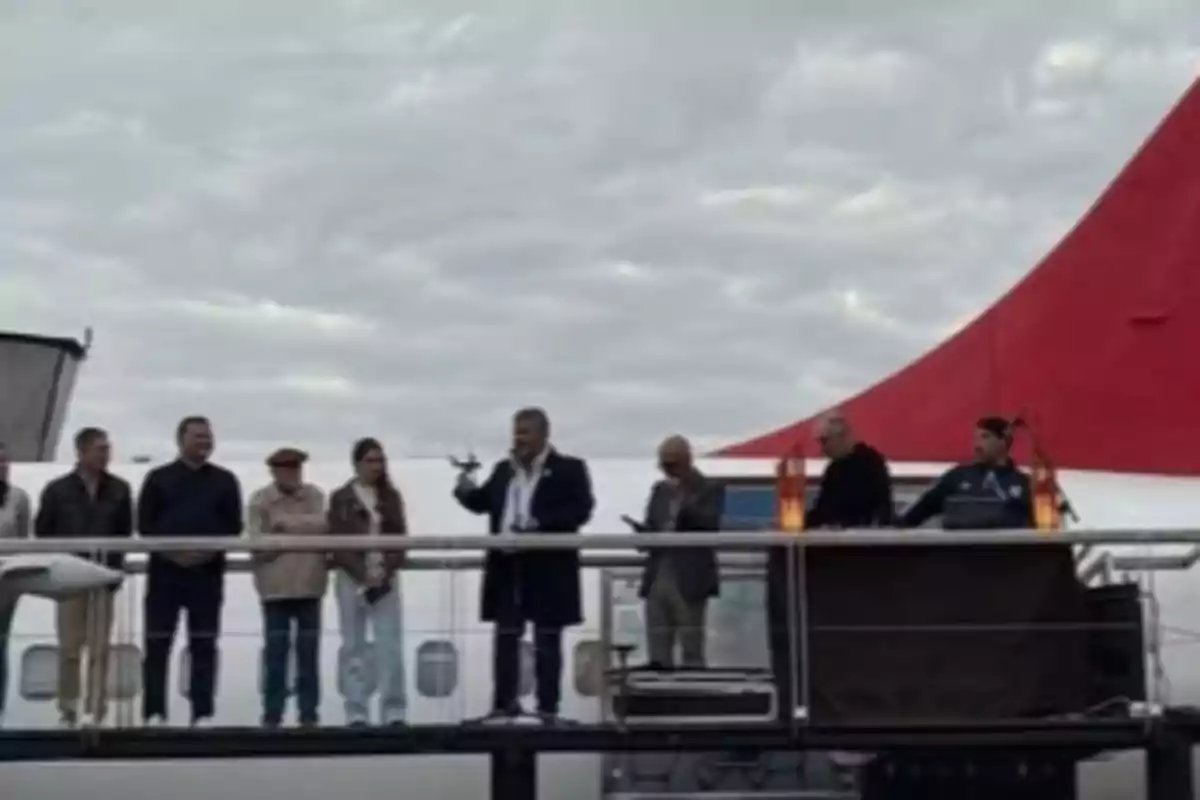 Several people are standing on a platform in front of a plane with a red tail, while one of them is holding an object and appears to be speaking to the audience. Several people are standing on a platform in front of a plane with a red tail, while one of them is holding an object and appears to be speaking to the audience.