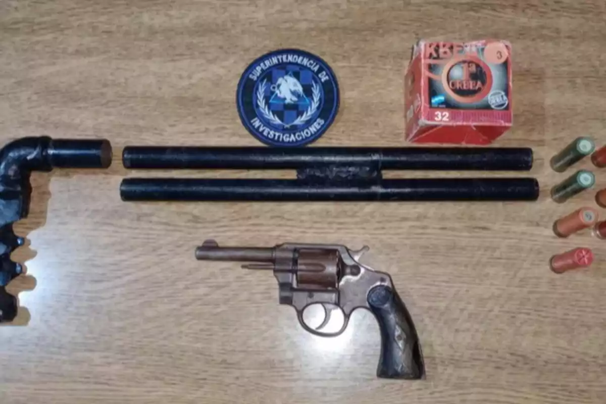 On a wooden table, there is a revolver, a homemade shotgun, cartridges, a box of ammunition, and a police patch. On a wooden table, there is a revolver, a homemade shotgun, cartridges, a box of ammunition, and a police patch.