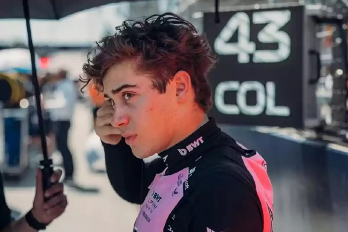 Young driver in a pink and black racing suit getting ready in the pit area with a sign in the background showing the number 43