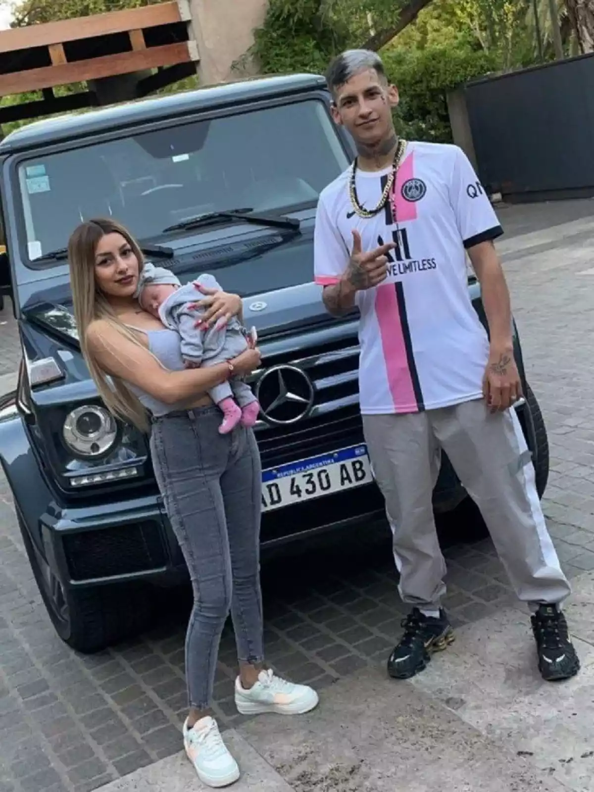 Una pareja joven con un bebé en brazos posa frente a una camioneta Mercedes-Benz negra estacionada en una calle de adoquines Una pareja joven con un bebé en brazos posa frente a una camioneta Mercedes-Benz negra estacionada en una calle de adoquines
