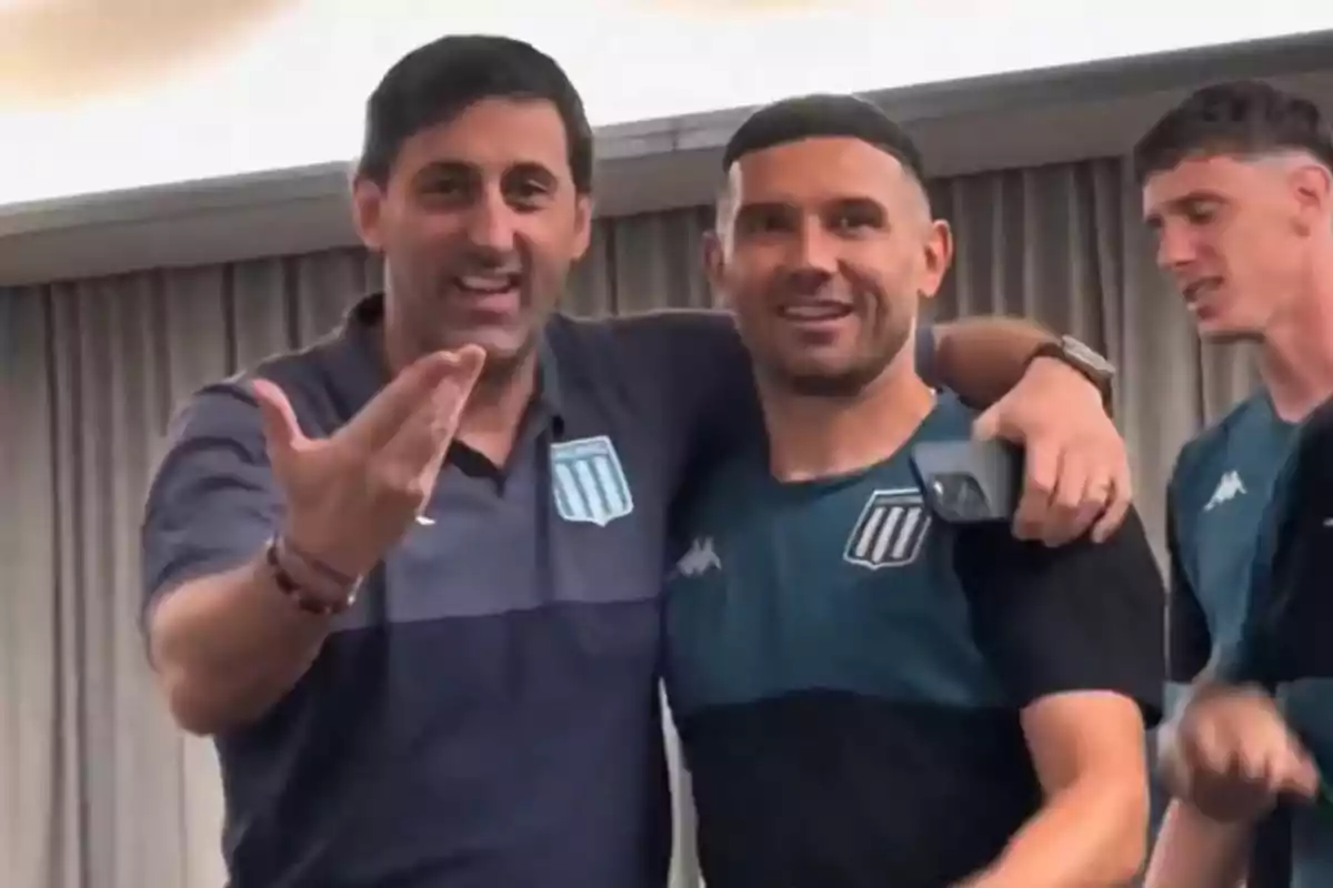 Two men smile and pose with their arms around each other wearing Racing Club sportswear while another man appears in the background. Two men smile and pose with their arms around each other wearing Racing Club sportswear while another man appears in the background.