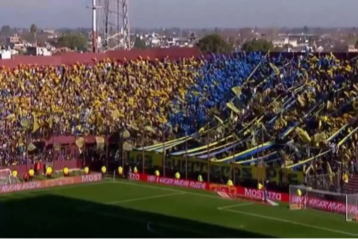 Tribuna de estadio llena de aficionados con banderas y globos amarillos y azules animando a su equipo de fútbol