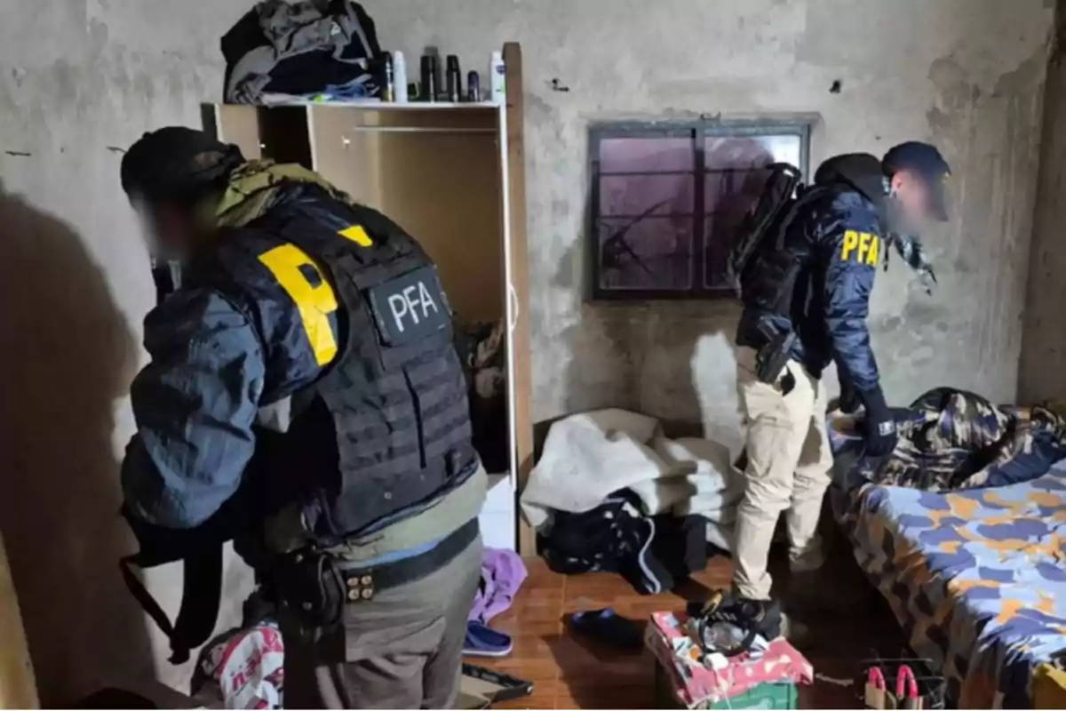 Dos agentes con chalecos de la PFA revisan una habitación desordenada con paredes desgastadas, una cama y un armario abierto.
