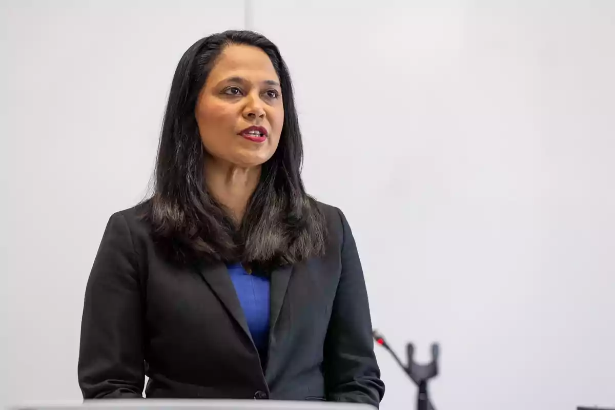 Woman with long dark hair wearing a black jacket and blue blouse speaking in front of a white background Woman with long dark hair wearing a black jacket and blue blouse speaking in front of a white background