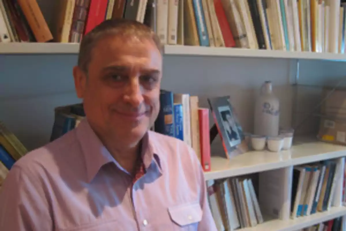 Hombre de cabello corto y camisa lila sonriendo frente a un estante lleno de libros