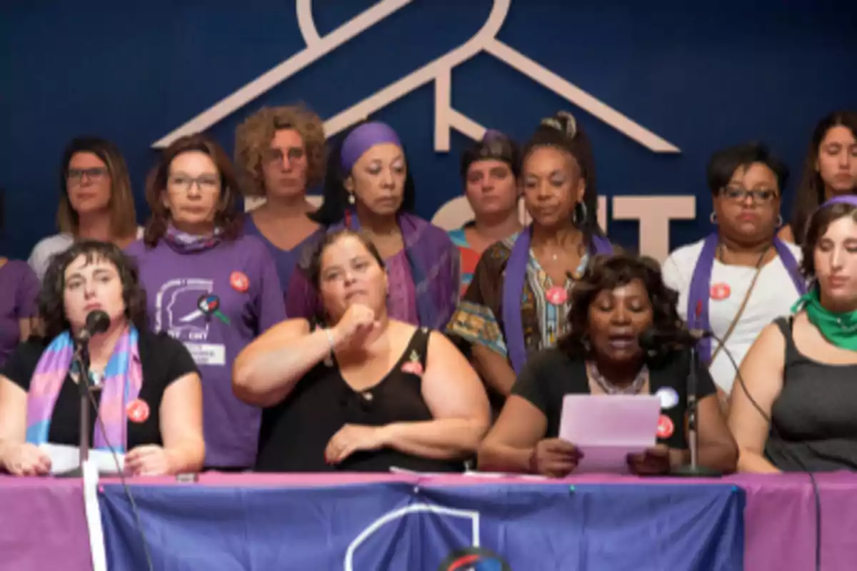 Grupo de mujeres reunidas en un escenario, algunas usando prendas moradas y pañuelos, mientras una de ellas lee un documento frente a un micrófono Grupo de mujeres reunidas en un escenario, algunas usando prendas moradas y pañuelos, mientras una de ellas lee un documento frente a un micrófono