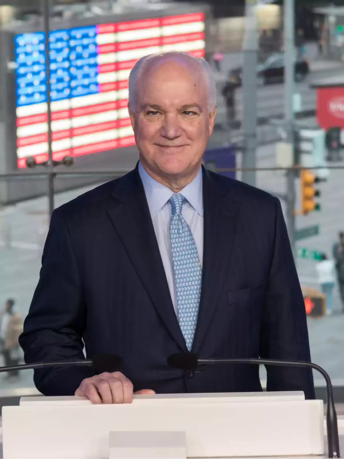 Gray-haired man in a dark suit and light blue tie standing in front of a podium with a blurred United States flag in the background Gray-haired man in a dark suit and light blue tie standing in front of a podium with a blurred United States flag in the background