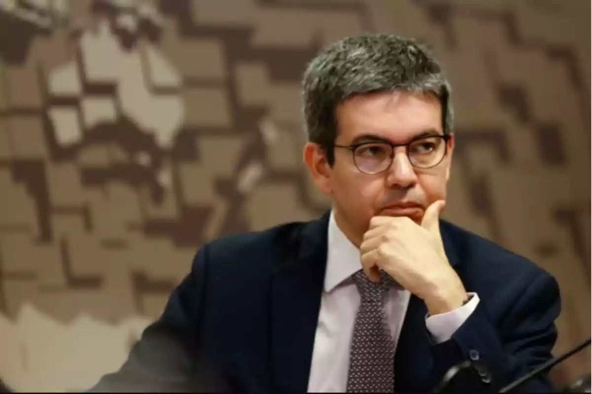 Gray-haired man with glasses and a thoughtful expression, dressed in a suit and tie, sitting in front of a background featuring a stylized world map design Gray-haired man with glasses and a thoughtful expression, dressed in a suit and tie, sitting in front of a background featuring a stylized world map design