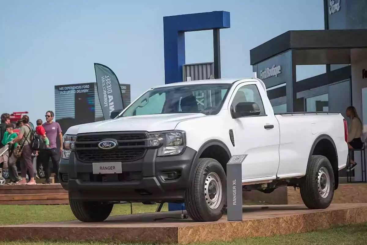 Camioneta Ford Ranger XL blanca exhibida en un evento al aire libre con personas alrededor y estructuras modernas de fondo Camioneta Ford Ranger XL blanca exhibida en un evento al aire libre con personas alrededor y estructuras modernas de fondo
