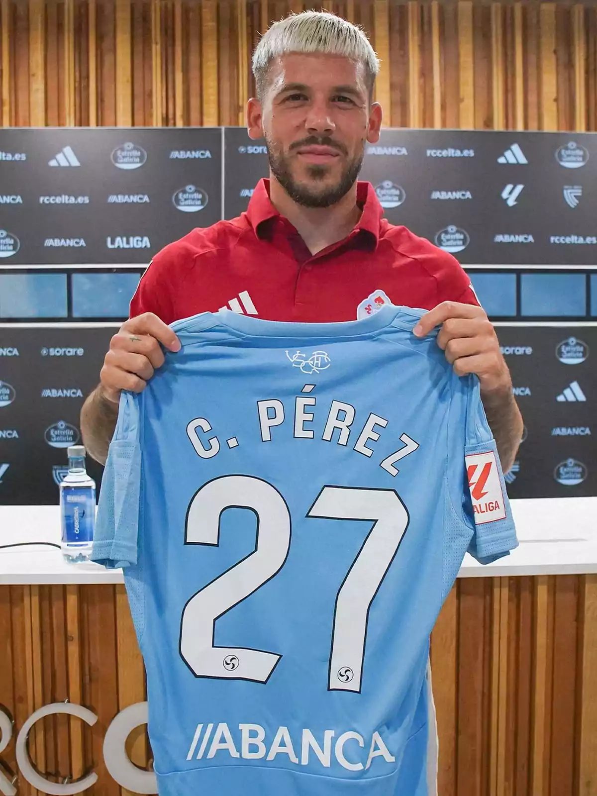 Blond-haired player holds a light blue Celta de Vigo jersey with the number 27 and the name C. Pérez in a press room. Blond-haired player holds a light blue Celta de Vigo jersey with the number 27 and the name C. Pérez in a press room.