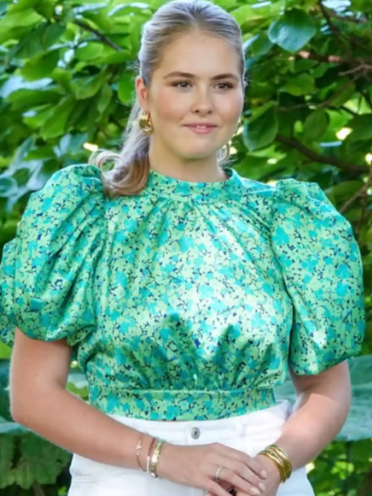 Young woman wearing a puff-sleeve blouse in green and blue tones posing outdoors in front of a background of green leaves Young woman wearing a puff-sleeve blouse in green and blue tones posing outdoors in front of a background of green leaves