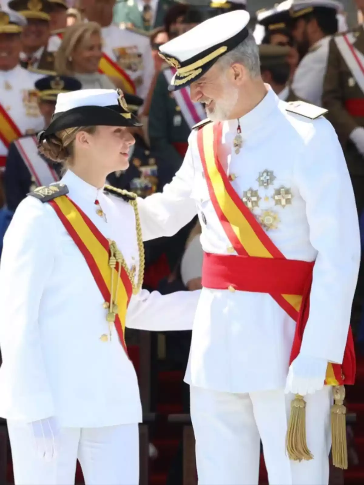 Two people dressed in white military uniforms with red and yellow sashes look at each other and smile at an official event surrounded by other uniformed individuals. Two people dressed in white military uniforms with red and yellow sashes look at each other and smile at an official event surrounded by other uniformed individuals.