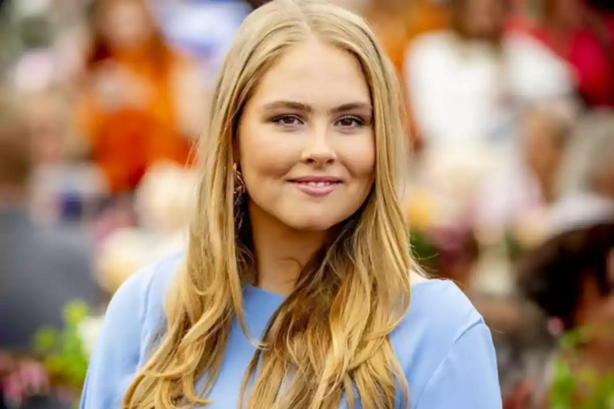 Young woman with long, loose blonde hair wearing a light blue blouse, smiling slightly outdoors with blurred people in the background