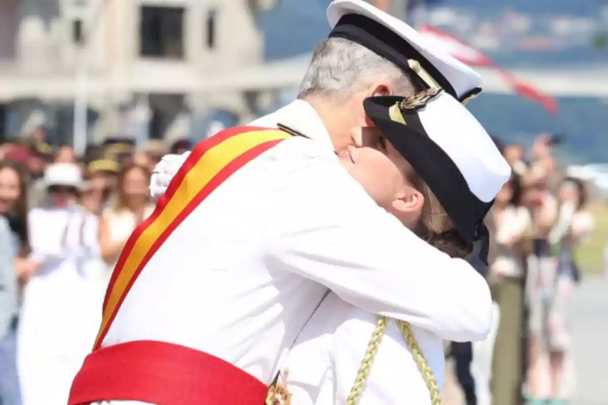 Two people dressed in white naval uniforms embrace at an official outdoor event while several people watch in the background.